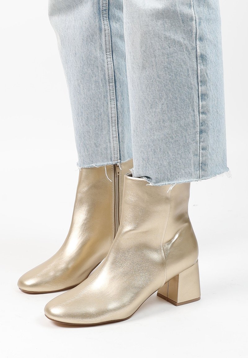 Bottines métalliques dorées avec une texture lisse, un bout pointu et un talon bloc. Portées avec un jean bleu clair usé qui s'arrête au-dessus de la cheville.