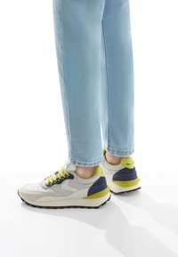 Sneakers multicolori con un upper beige, grigio e blu navy, lacci gialli luminosi e suola nera testurizzata, abbinati a jeans azzurri chiari.