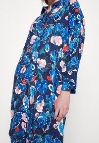 Abito camicia floreale in blu navy con vivaci motivi blu, rosa e verdi. Presenta un colletto, chiusura con bottoni e maniche lunghe. Tessuto morbido.