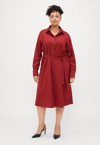 Vestido camiseiro bordô com mangas longas, frente abotoada e cintura cinto. Feito de tecido suave, com um brilho suave e fendas laterais.