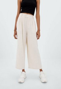 Wijdvallende broek in lichtbeige stof met plooien, voorzien van twee knopen in de taille. Gecombineerd met witte sneakers.