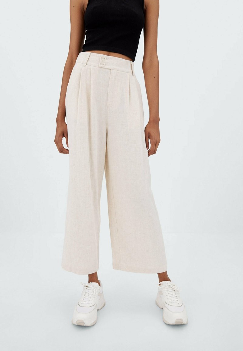 Wijdvallende broek in lichtbeige stof met plooien, voorzien van twee knopen in de taille. Gecombineerd met witte sneakers.