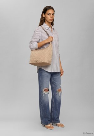 Γυναίκα με ριγέ πουκάμισο, σκισμένο μπλε τζιν και σανδάλια κρατώντας μπεζ καπιτονέ τσάντα ώμου μπροστά σε γκρι φόντο.