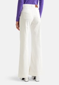 Vita vida byxor i bomull med hög midja, med två bakfickor och en brun läderlogga vid midjan.