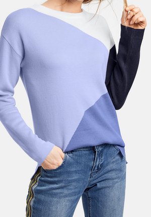 Person trägt einen Colorblock-Pullover in Weiß, Lavendel und Marineblau mit blauen Jeans mit goldenen und schwarzen Seitenstreifen, Hand in der Tasche.