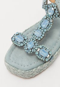 Sandale en daim bleu clair avec un ornement de gemmes délicatement conçu, comportant différentes nuances de pierres bleues et une plateforme en jute tressé.