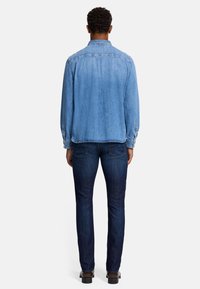 Lichtblauwe spijkerblouse met lange mouwen, geknoopte manchetten en een ontspannen pasvorm, gedragen met donkerblauwe rechte jeans en bruine schoenen.