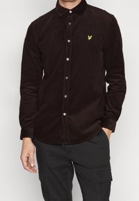 Camisa de pana marrón con botón y cuello puntiagudo, que presenta un pequeño logotipo amarillo en el lado izquierdo del pecho y botones blancos en la parte delantera.