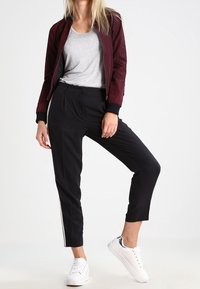 Burgundy bomber jacket over a gray t-shirt, paired with black cropped pants featuring side stripes, and white sneakers. Smooth fabrics.