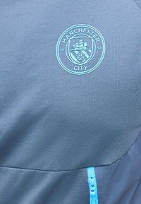 Λογότυπο της Manchester City σε ανοιχτό γαλάζιο σε σκουρό μπλε ύφασμα πάνω από ένα ανοιχτό γαλάζιο φερμουάρ σε μπουφάν ή αθλητικό ένδυμα.