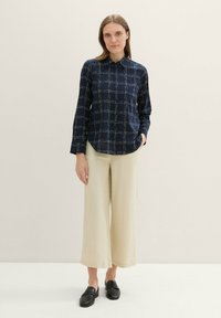 Chemise bleu marine à boutons avec un motif de carreaux blancs, à manches longues et col, associée à un pantalon large beige clair et des mocassins noirs.