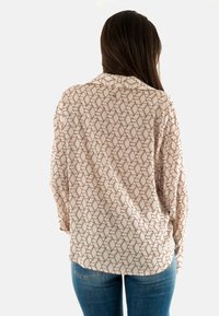 Camisa de manga larga en beige claro con un patrón geométrico marrón, que presenta un cuello y un corte relajado, combinada con vaqueros azules.
