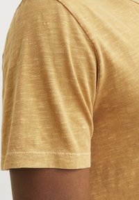Camiseta de manga corta en un color mostaza claro, con una tela texturizada que presenta un patrón sutil y una costura ordenada a lo largo del dobladillo.