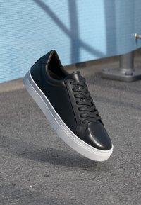 Black synthetic leather sneaker with a smooth finish, white rubber sole, and flat black laces. Features rounded toe and minimalistic design.