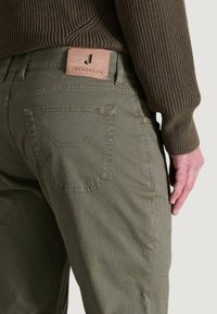 Pantalones de algodón verde oliva con un bolsillo trasero con costura triangular y una etiqueta de cuero de marca en la cintura. Textura sutil.