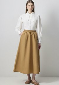 Blusa blanca de cuello con botones, combinada con una falda larga de cintura elástica en color marrón sólido. La modelo está de pie con las manos en los bolsillos, usando zapatos planos.
