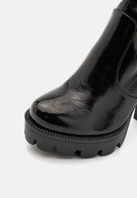 Musta nilkkuri, jossa on kiiltävän lakattu pinta, paksu kumipohja, jossa on teksturoitu reuna, sekä kärjeltään terävä muoto.