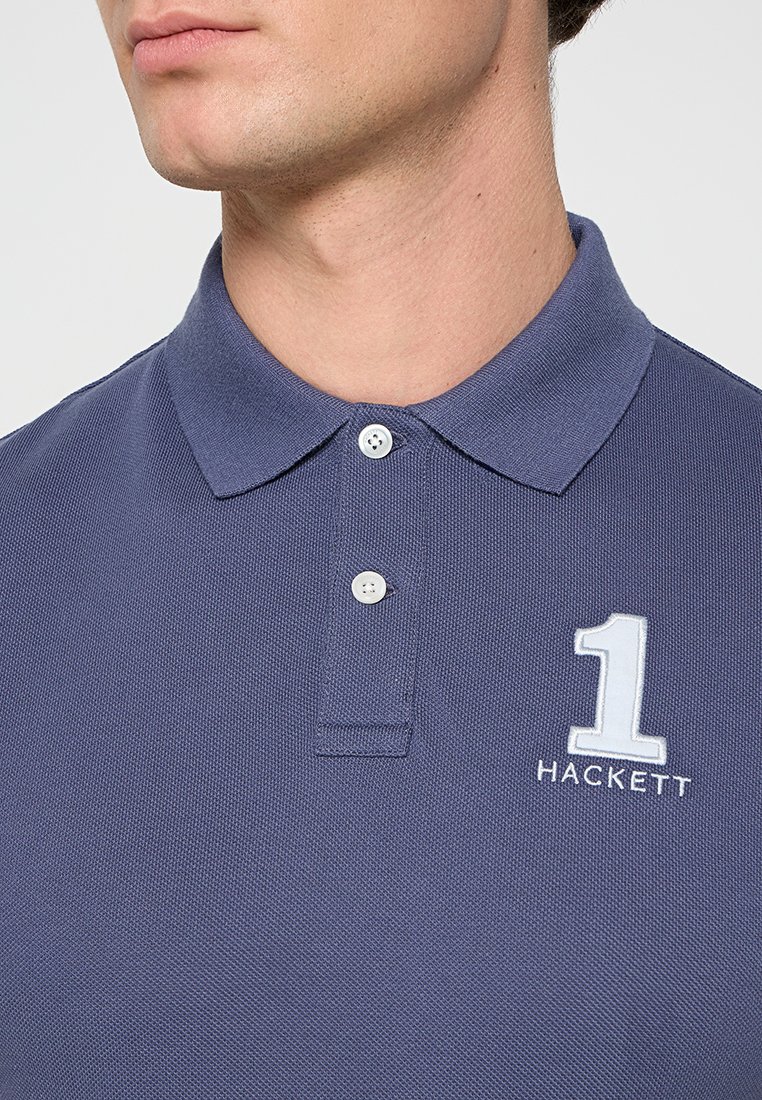 Homme portant un polo bleu à col avec deux boutons blancs et un logo blanc en forme de "1" au-dessus de "HACKETT" sur la poitrine.