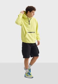 Felpa gialla neon con tasca frontale e dettagli neri, abbinata a pantaloni cargo neri e sneakers colorate con calzini a fantasia.