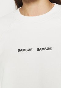 Sudadera blanca de cuello redondo hecha de algodón con el texto negro "SAMSKU" impreso dos veces en el frente. Cuenta con un cuello ribeteado y una textura suave.