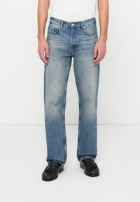 Man wearing light blue straight-leg jeans and black tassel loafers against white background, hands resting by sides.