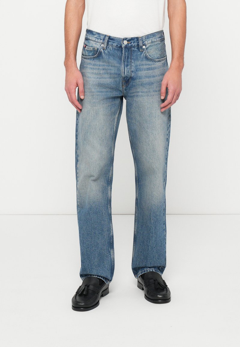 Man wearing light blue straight-leg jeans and black tassel loafers against white background, hands resting by sides.