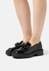 Musta lakattu loafers-kengät paksulla pohjalla, joissa on koristeellinen ketju etuosassa ja sileä pinta. Sopii rentoon käyttöön.
