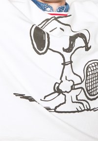 Sudadera blanca con un contorno negro de un perro de dibujos animados sosteniendo una raqueta de tenis, con un cuello de mezclilla visible debajo.