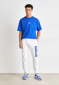 Blaues Baumwoll-T-Shirt mit kurzen Ärmeln, weiße Fleece-Jogginghosen mit blauem "HUGO"-Logo und blau-weiße Sneaker mit dicker Sohle.