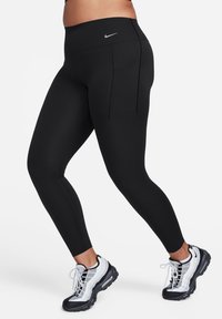 Svarte leggings laget av elastisk stoff har høy midje og to baklommer. Kombinert med svart-hvite treningssko.
