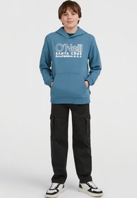 Gutt som står med hendene i lommene, iført blå O'Neill hettegenser, svarte cargo-bukser og hvite joggesko, mot en ensfarget bakgrunn.