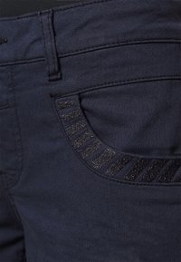 Jeans de mezclilla azul oscuro con un bolsillo texturizado que presenta franjas brillantes y mate alternas. Vista en primer plano de la tela y los detalles.