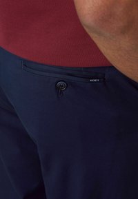 Marineblauwe broeken met een gladde textuur, voorzien van een frontzak met een zwarte knoop en subtiele "HACKETT" branding op de tailleband.