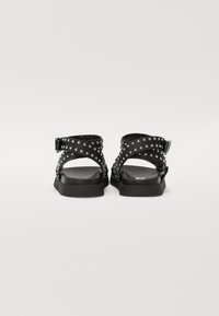 Sorte sandaler med naglede stropper, som har en flad sål og justerbare ankel-lukninger. Enkel, fleksibel design.