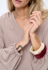 Beige blouse met poffende mouwen; een wit en bordeauxrood harig armbandje omgeeft de pols. Zwarte zigzag tatoeage op de onderarm.