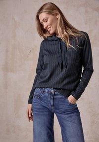 Marine gestreiftes Langarmoberteil mit Kordelzug-Halslinie, kombiniert mit hellblauen weiten Jeans. Sanfte Textur, lässige Passform und sichtbare Taschen.