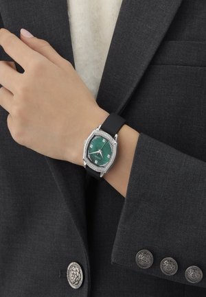 Octagonal watch with a green dial, silver accents, diamond-like embellishments, and a black rubber strap, worn on a hand with a gray blazer.