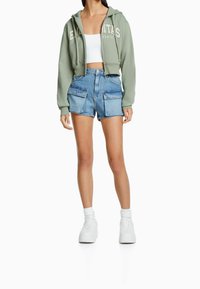 Mujer vistiendo una sudadera corta con cremallera en verde salvia, una camiseta corta blanca, pantalones cortos de mezclilla de tiro alto con bolsillos en la parte delantera, calcetines blancos y zapatillas de plataforma.