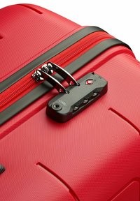 Valise rigide rouge avec une surface texturée, zip noir, tirettes de zip métalliques doubles et une serrure approuvée par la TSA avec un panneau de contrôle.