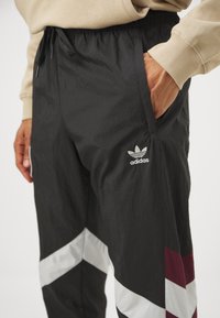 Pantalones deportivos negros con un acabado texturizado, que presentan un diseño de flecha blanco y burdeos y un logo de Adidas en el muslo izquierdo.