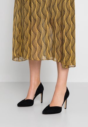 Chaussures à talons hauts en suède noir avec un bout pointu et un design épuré, assorties à une jupe jaune à motifs présentant un design de vagues.