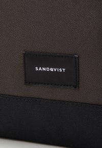 Mørk grå tekstilbag med et sort stoffetikett som leser "SANDQVIST." Har en teksturert overflate og kontrasterende svarte detaljer.
