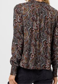 Langärmlige Bluse mit Paisley-Muster in Schwarz, Rot und Gold. Verfügt über einen gerafften Ausschnitt und Bündchen, gefertigt aus einem strukturierten Stoff.