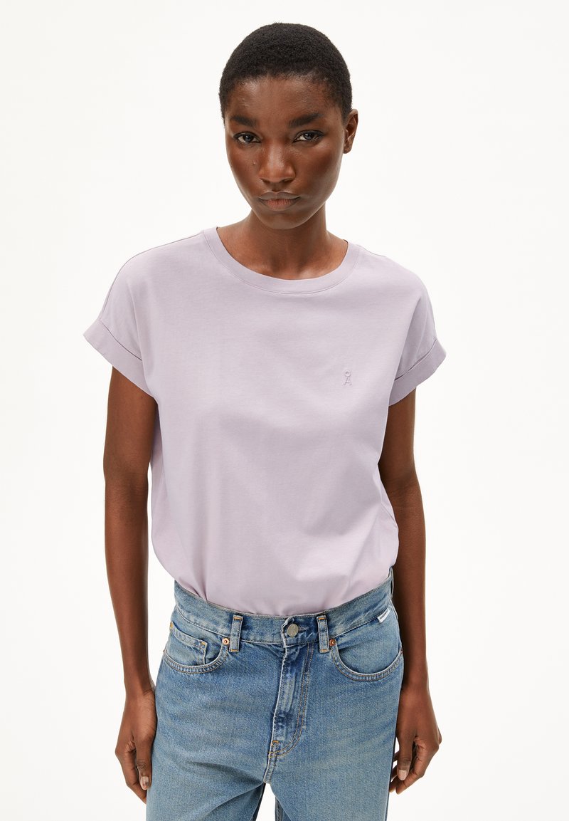 Lila Baumwoll-T-Shirt mit aufgerollten Ärmeln und einem kleinen gestickten Logo auf der Brust, kombiniert mit hellblauen Jeans.