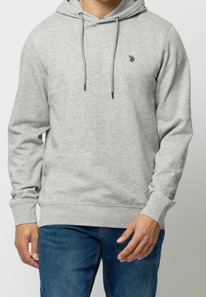 Mann trägt hellgrauen Kapuzenpullover mit kleinem Logo auf der linken Brust und blaue Jeans, steht mit entspannten Armen.