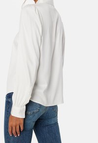 Blouse blanche à manches longues avec une coupe décontractée, présentant une texture délicate et des poignets boutonnés, portée avec un jean en denim bleu.