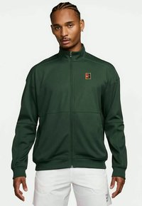 Junger Mann mit geflochtenem Haar, der eine dunkelgrüne Zip-Jacke und weiße Shorts trägt und vor einem schlichten hellen Hintergrund steht.