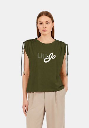 Donna che indossa una canotta verde oliva senza maniche con coulisse e logo "Liu Jo", abbinata a pantaloni beige, in piedi davanti a uno sfondo semplice.