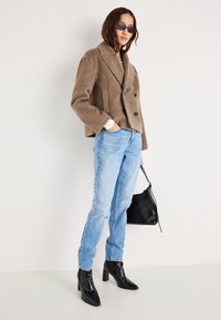 Chaqueta cruzada de mezcla de lana marrón con solapas anchas, vaqueros de pierna recta azul claro y botines negros de punta afilada. Sostiene un bolso negro.