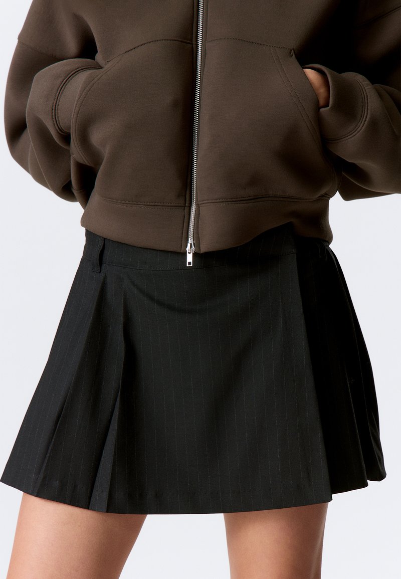 Mustat raidalliset hameet, joissa on laskokset, yhdistettynä ruskeaan vetoketjulliseen collegepaitaan. Kangas näyttää sileältä ja hieman tekstuurilta.